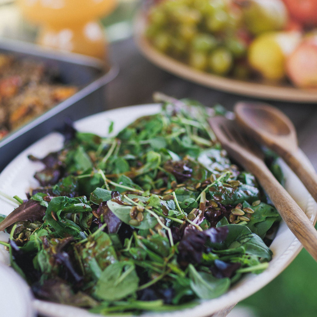 Green salad in a white bowl with wooden utensils on a blurred background of fruits.